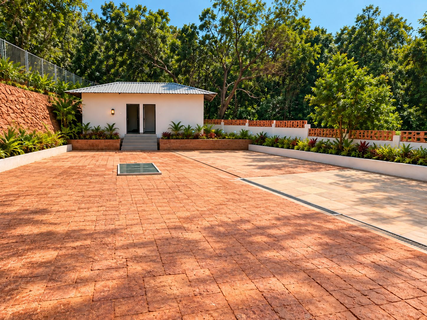 A paved courtyard with a small building, surrounded by lush greenery and a stone wall.