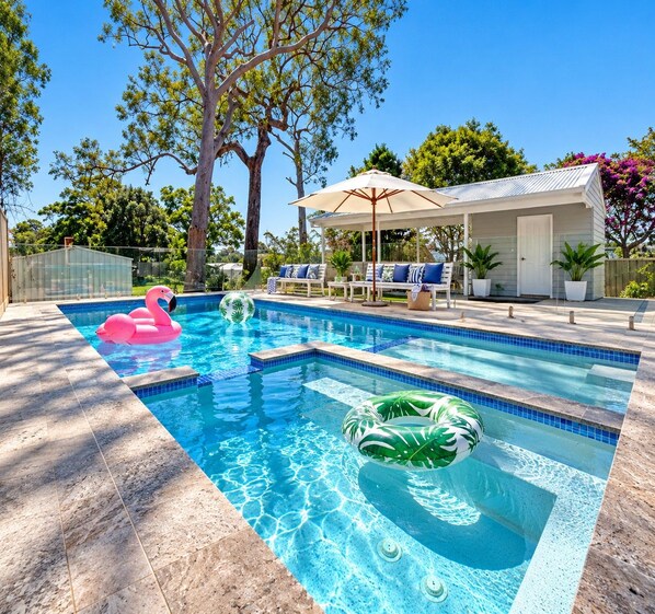 A swimming pool with a lounge area, a covered seating area, and a view of trees and a house.