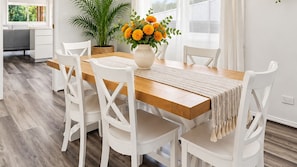 A dining area with a wooden table, white chairs, a vase with flowers, and a potted plant. - Fairways at the Bay - Heated Pool and Green View (Nelson Bay)
