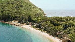 An aerial view of a coastal area with a beach, dense forest, and a clear view of the sea.
