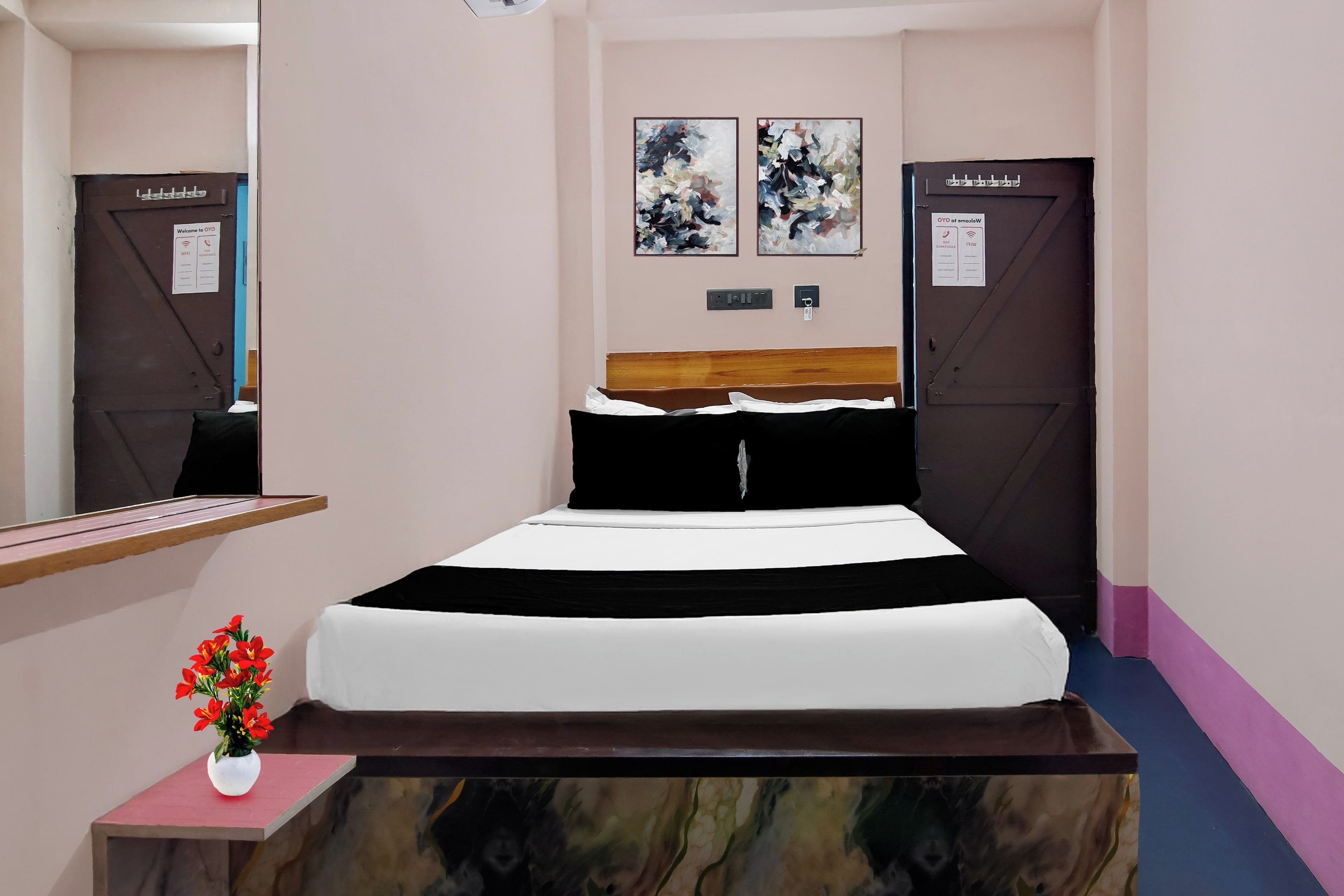 A hotel room with a bed, a bedside table with a vase of flowers, a wall-mounted power outlet, and two doors with signs.