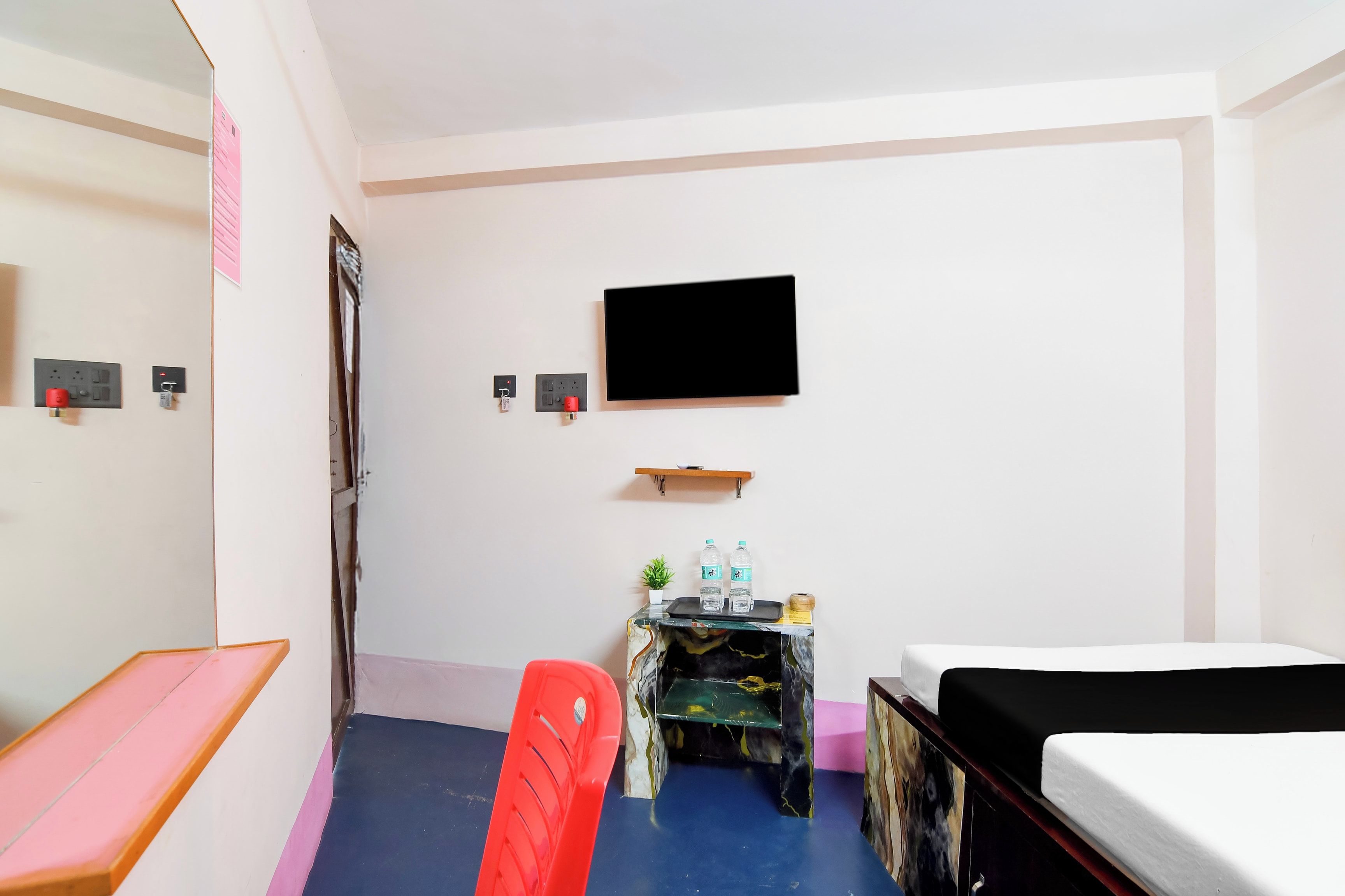 Una habitación de hotel con cama, escritorio con silla, televisor en la pared y una mesita con una planta y botellas de agua.