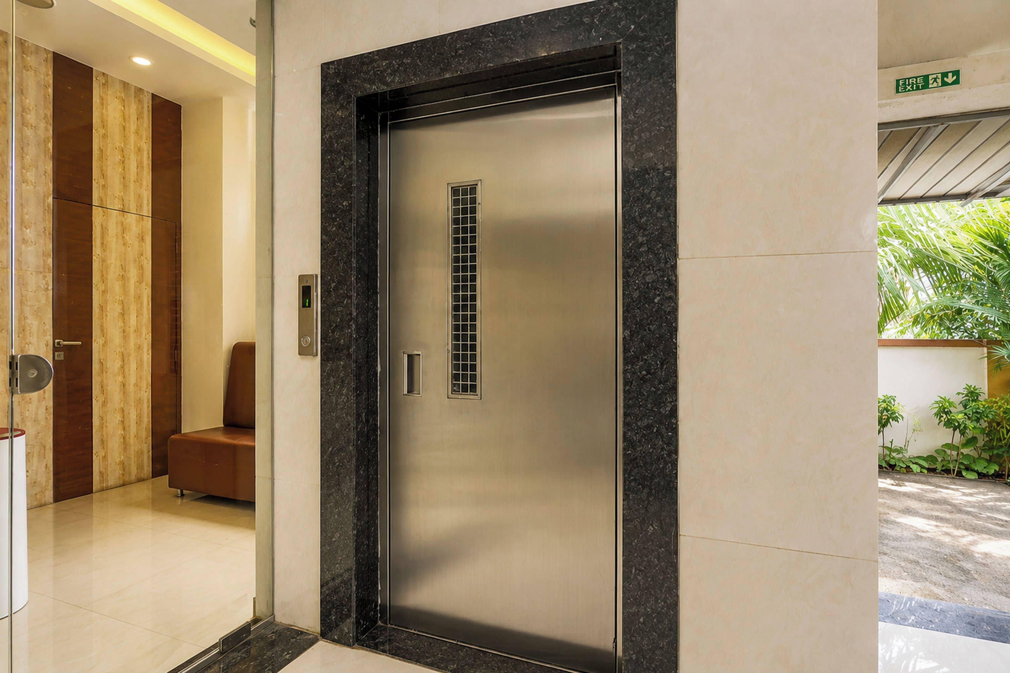 A modern elevator with a metallic finish and a glass panel, located in a hallway with a wooden panel wall and a bench.