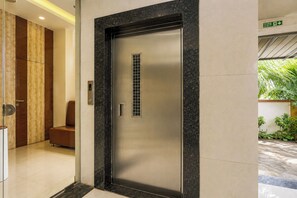 Un ascensor moderno con detalles metálicos y panel de vidrio, situado en un pasillo con revestimiento de madera y un banco.