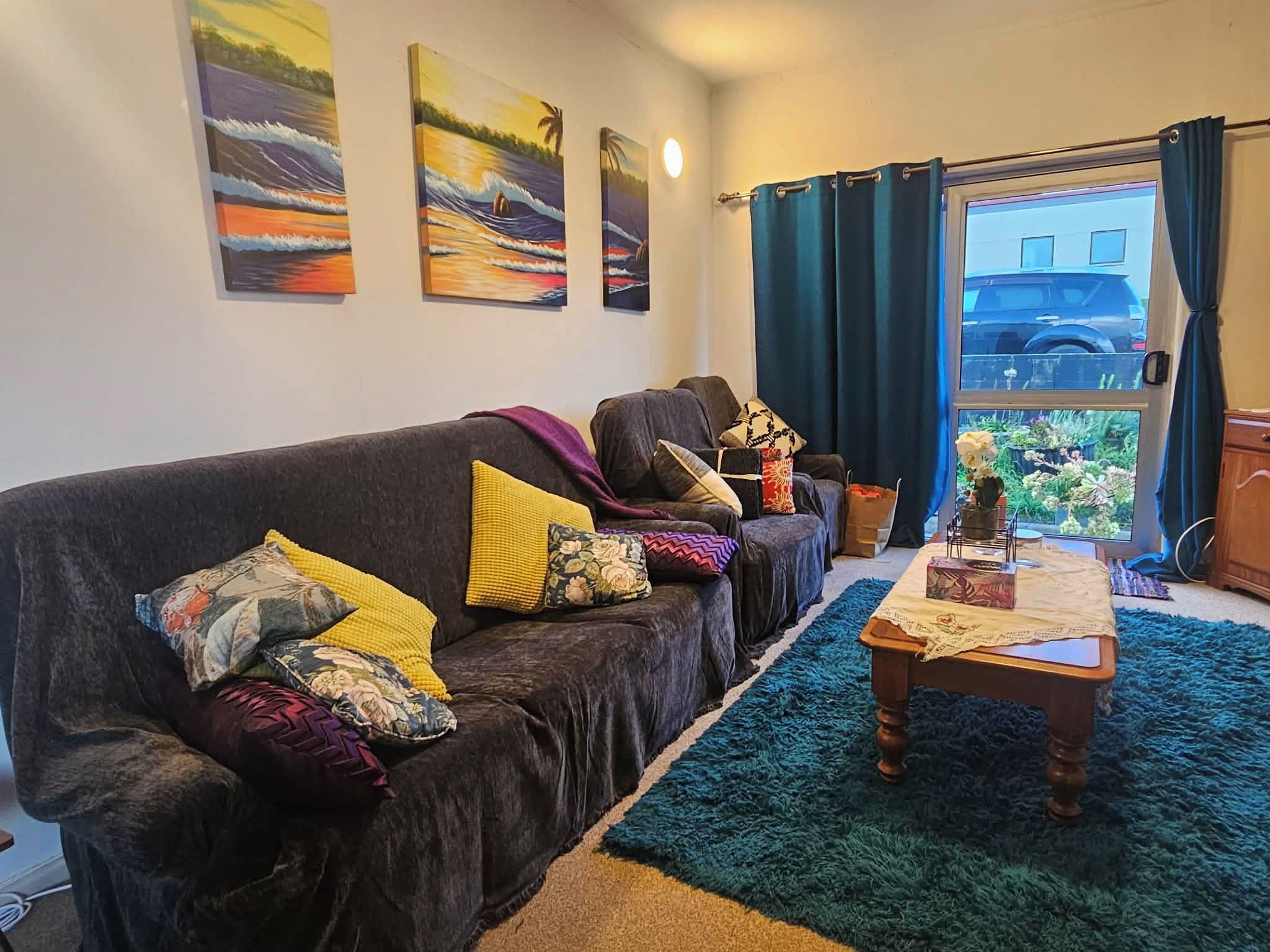 A living room with a dark-colored sofa, multiple cushions, a wooden coffee table, and a view of the outside through a window with curtains.