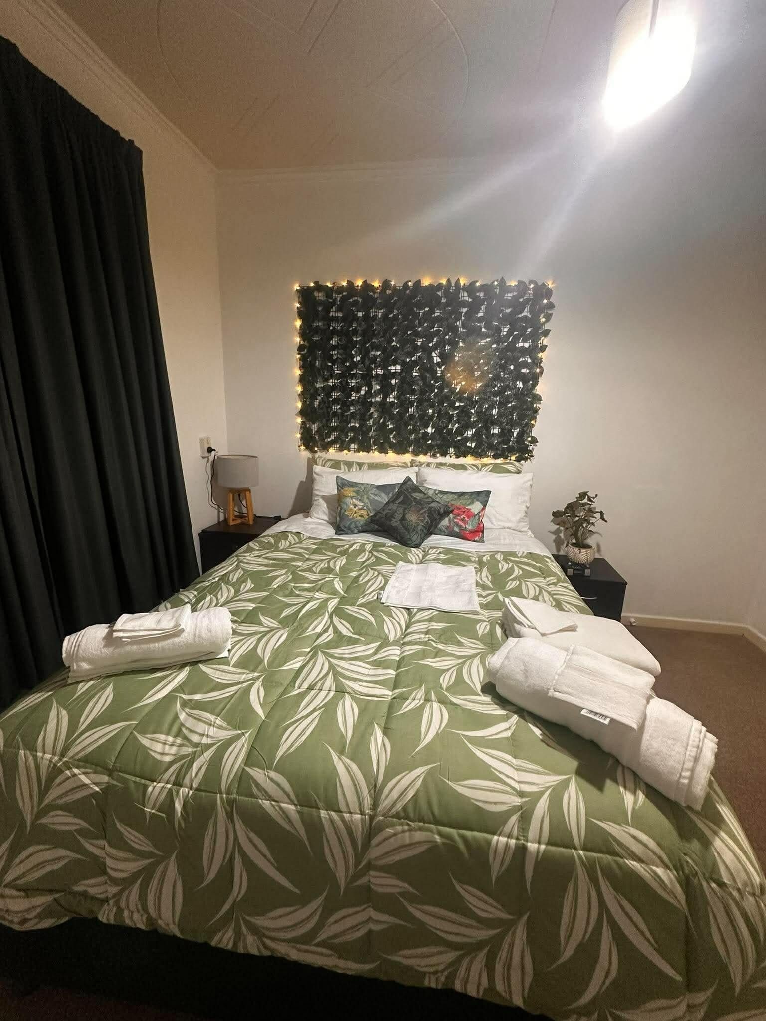 A neatly made bed with a green leaf-patterned bedspread, a headboard with string lights, and a nightstand with a lamp and a small plant.
