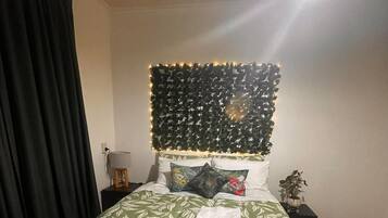 A neatly made bed with a green leaf-patterned bedspread, a headboard with string lights, and a nightstand with a lamp and a small plant.
