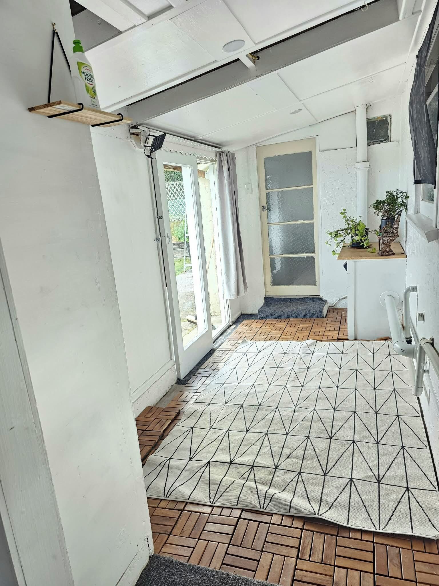 Un couloir étroit doté d’un sol à motifs, une porte donnant sur un balcon et un petit coin cuisine agrémenté d’une plante.