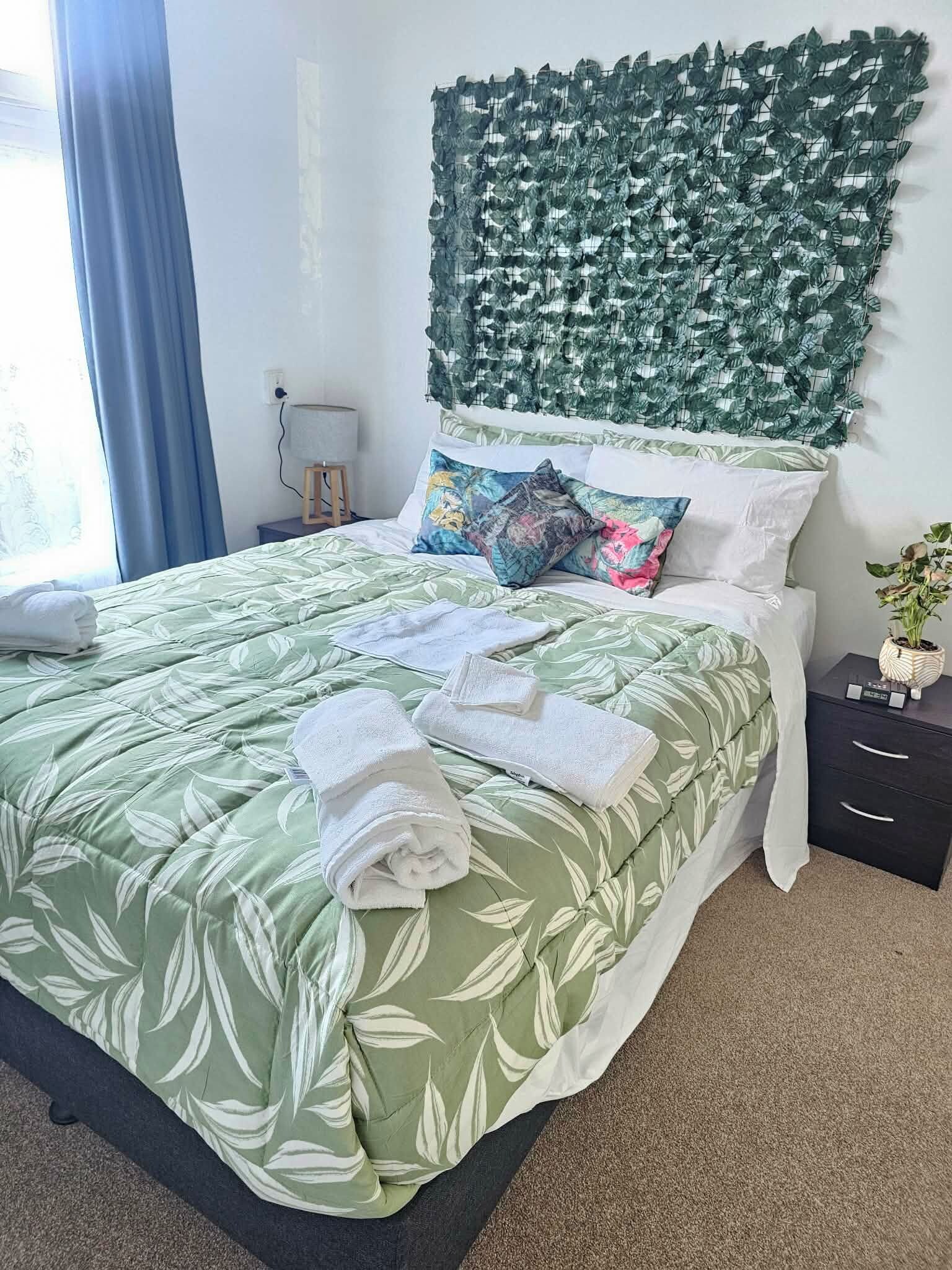 Una cama bien hecha con una colcha de estampado de hojas, una planta en la pared y una mesita de noche con una lámpara y una planta pequeña en maceta.