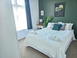 A neatly made bed with white linens and towels, a bedside table with a lamp, a framed floral artwork, and a large window with curtains.