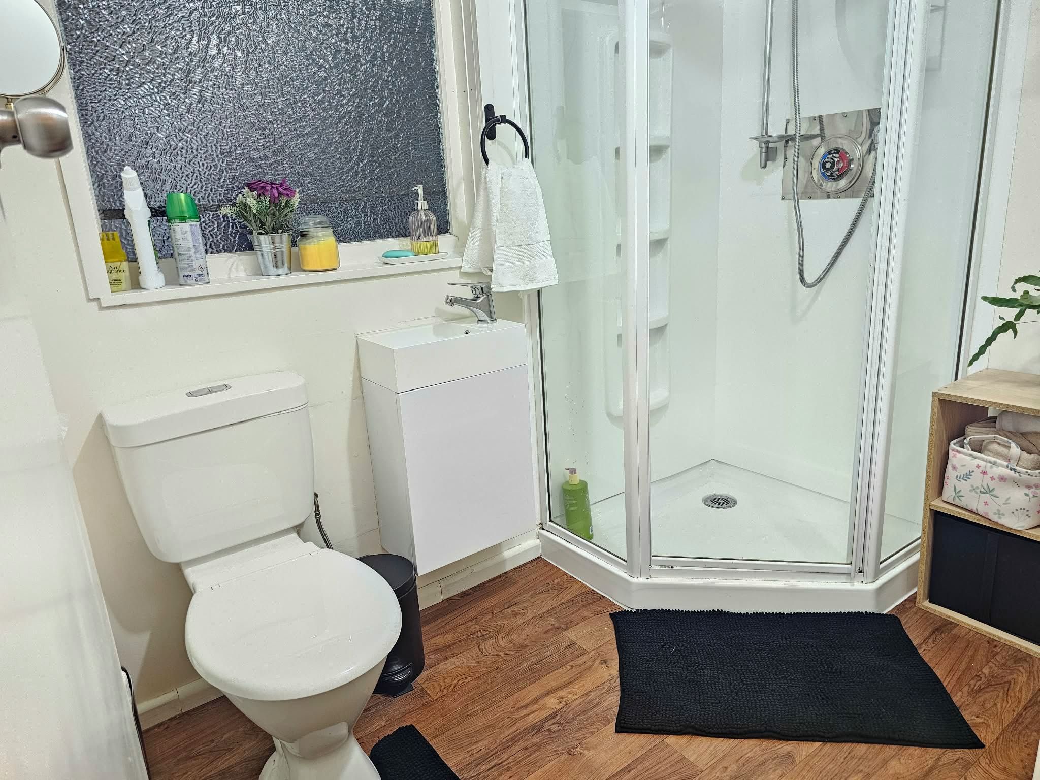 Une salle de bain équipée de toilettes, d’un lavabo, d’une douche et d’une petite étagère en bois où sont posées des serviettes.