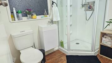 Une salle de bain équipée de toilettes, d’un lavabo, d’une douche et d’une petite étagère en bois où sont posées des serviettes.