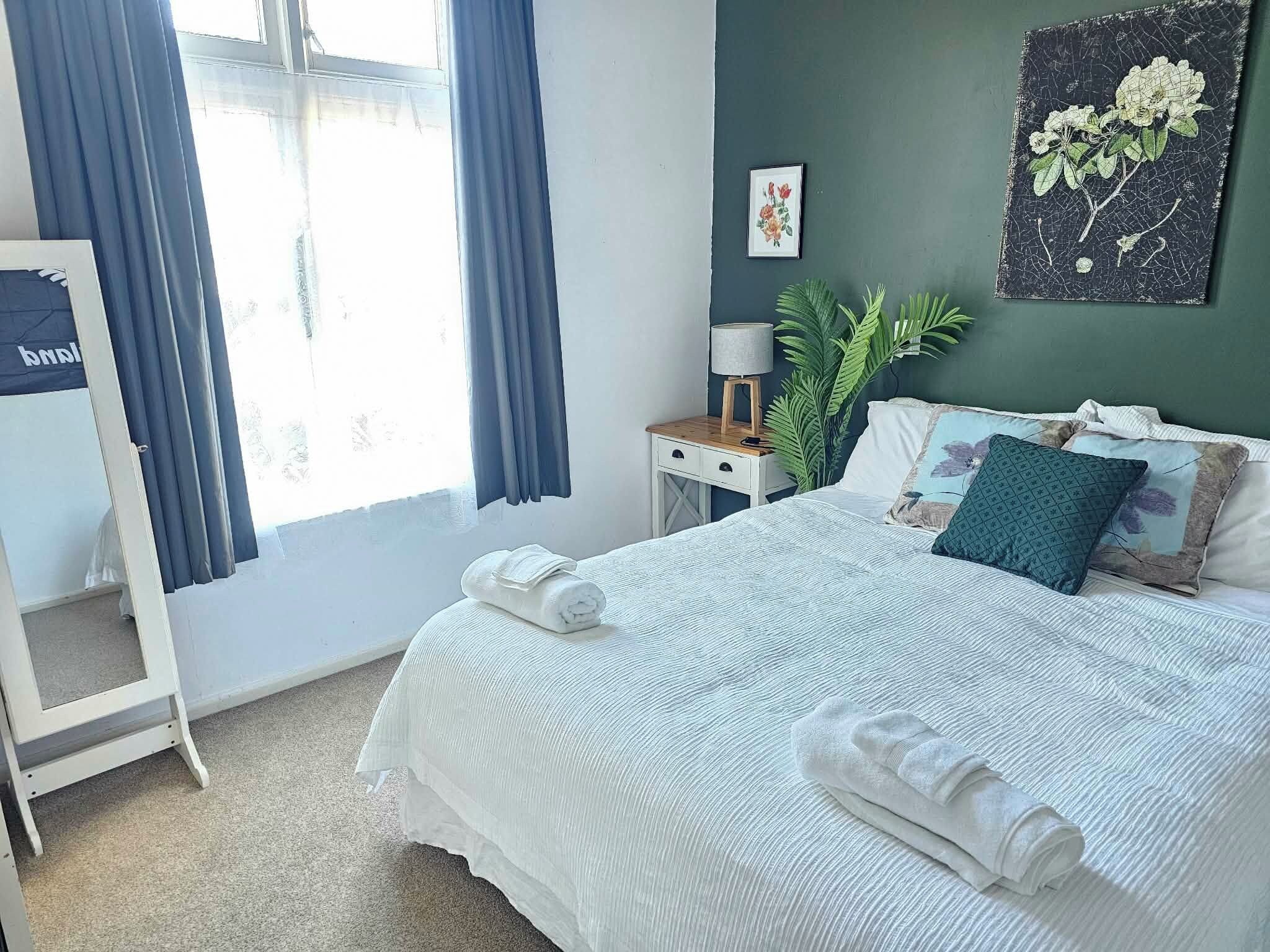 Ein ordentlich bezogenes Bett mit weißen Leinen und gerollten Handtüchern, ein Nachttisch mit Lampe, ein Topfpflanze und ein gerahmtes Pflanzenbild an der Wand.