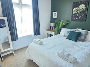 A neatly made bed with white linens and rolled towels, a bedside table with a lamp, a potted plant, and a framed botanical illustration on the wall.