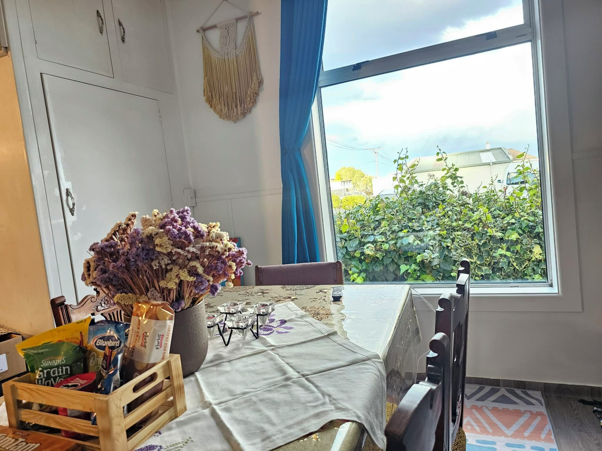 A room with a table set for breakfast, a window with a view of greenery, and a macrame hanging on the wall.