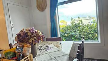 A room with a table set for breakfast, a window with a view of greenery, and a macrame hanging on the wall.