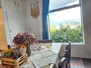 A room with a table set for breakfast, a window with a view of greenery, and a macrame hanging on the wall.