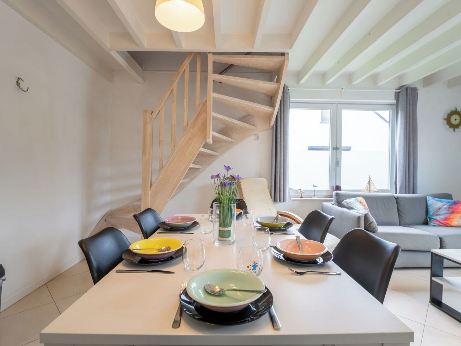 A modern dining area with a table set for four, a staircase, and a living room with a sofa and a view through a large window.