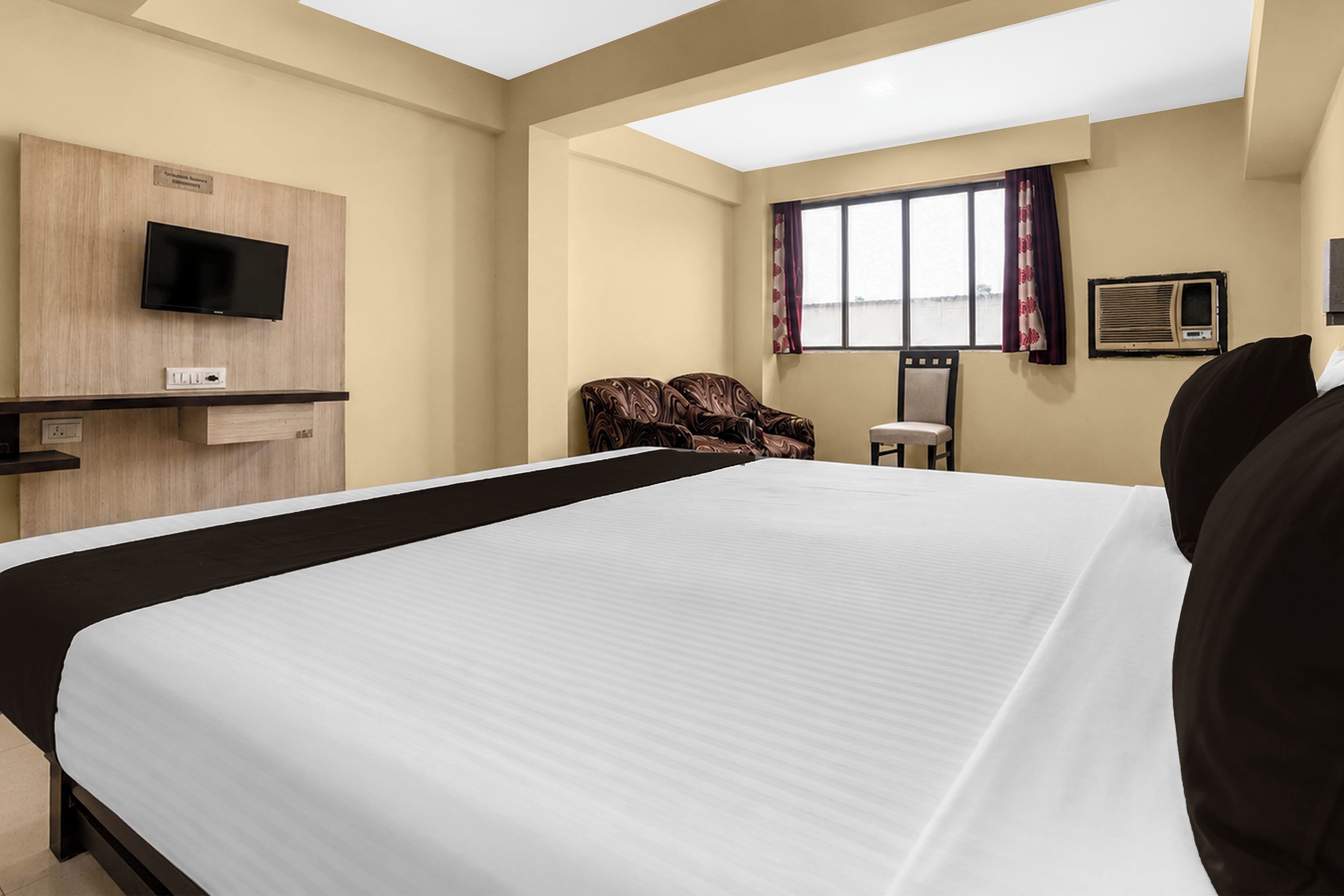 Habitación de hotel con una cama grande, televisor montado en la pared, una silla y un ventanal con cortinas.
