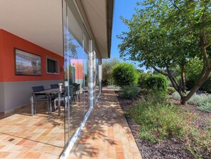 Eine Terrasse im Freien mit Glasveranda, einem Tisch mit Stühlen und einem Garten mit verschiedenen Pflanzen und Blumen.