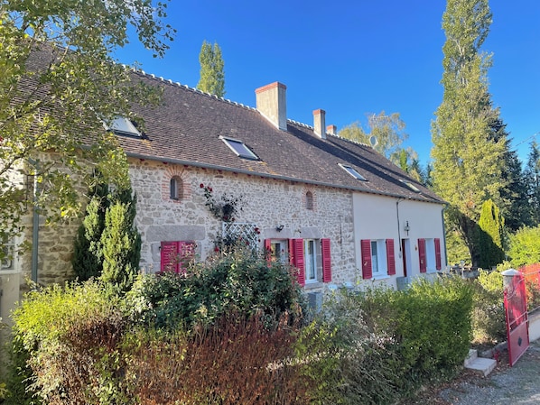 A stone house with a tiled roof, red shutters, and a garden in front.