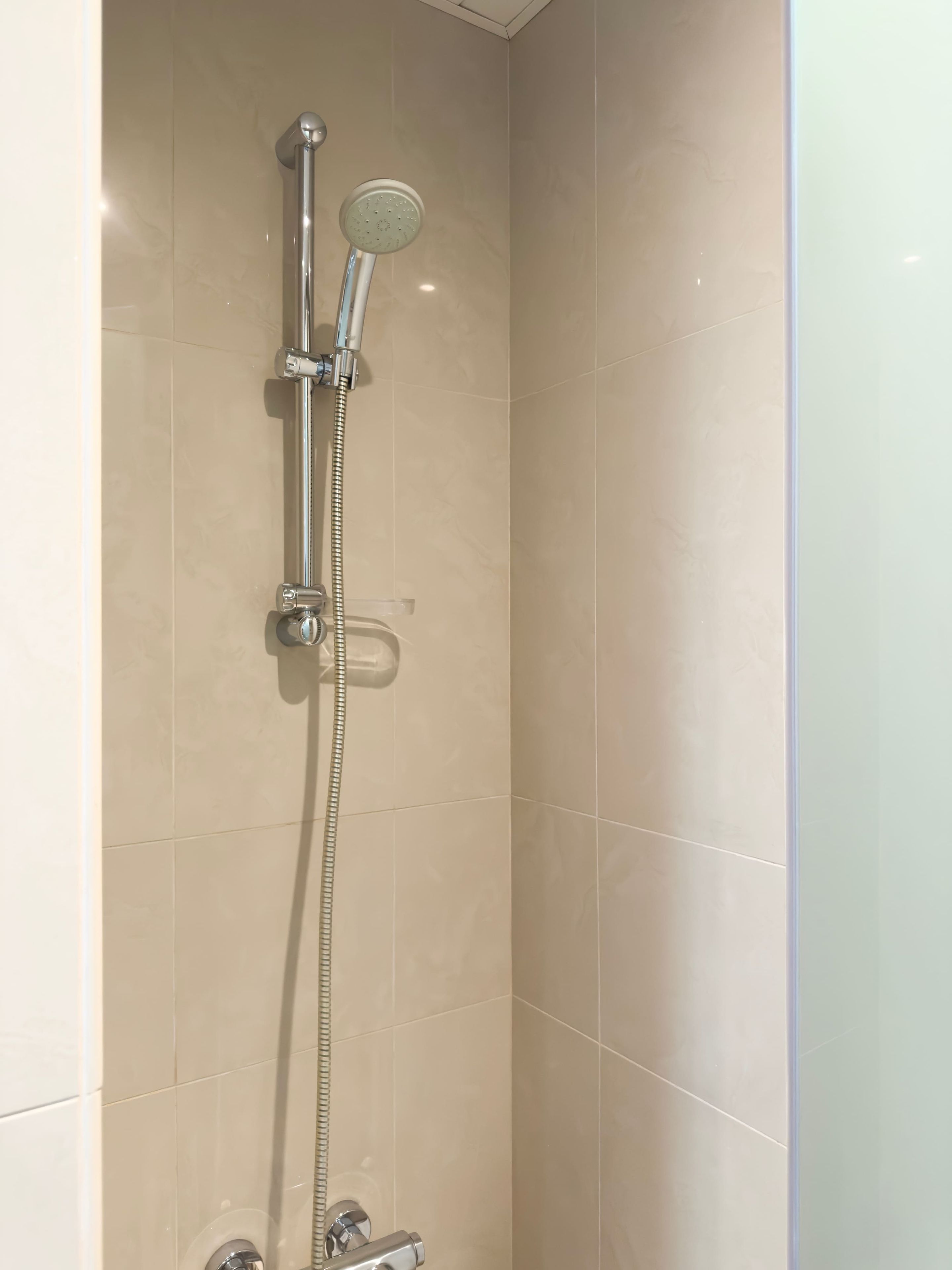 Une douche équipée d’un pommeau de douche à main et d’un pommeau de douche fixe fixé au mur, tous deux dotés de finitions chromées, dans une salle de bain aux murs recouverts de carreaux beiges.