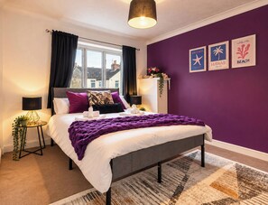 A bedroom with a bed, a window with curtains, a wall with framed pictures, and a bedside table with a lamp. - Westend Parade (Gloucester)