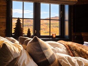 A bedroom with a large window, a bed with white and brown bedding, and a view of a forested landscape. - Modern Alpine Living in Ustaoset (Hol)