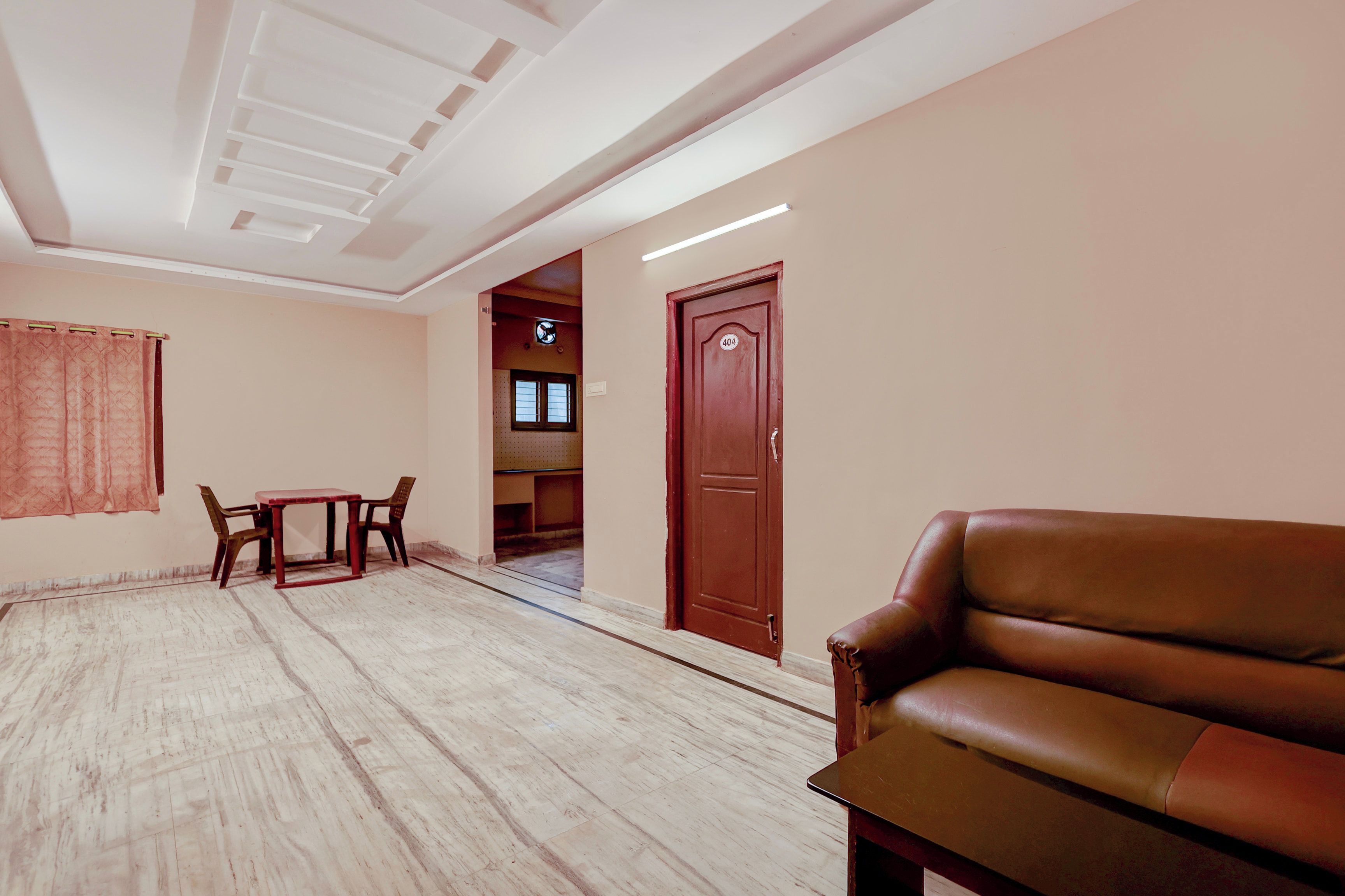 Une chambre d’hôtel avec un plancher en bois, un canapé en cuir marron, une petite table avec deux chaises, une porte avec une plaque numérotée et une fenêtre avec des stores.