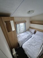 Una habitación pequeña con dos camas, una ventana con cortinas, una lámpara en el techo y un estante de madera.