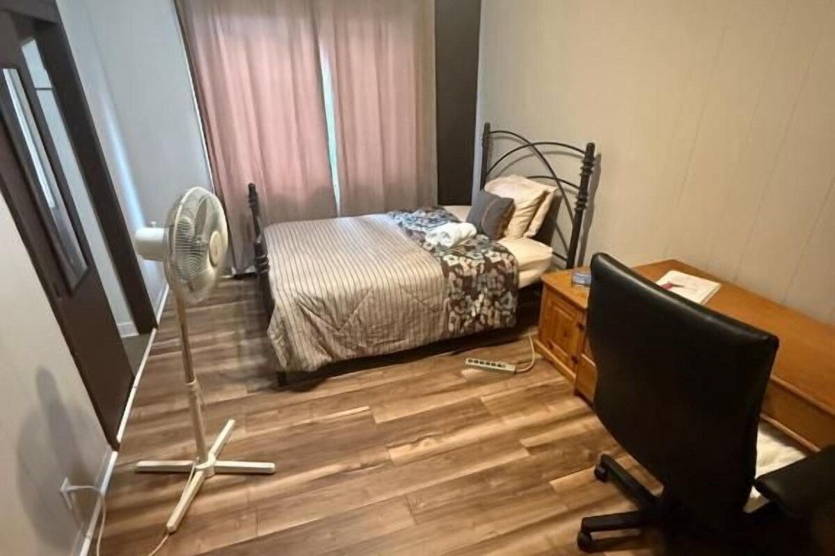 A bedroom with a bed, a desk, a chair, a fan, and a window with curtains.