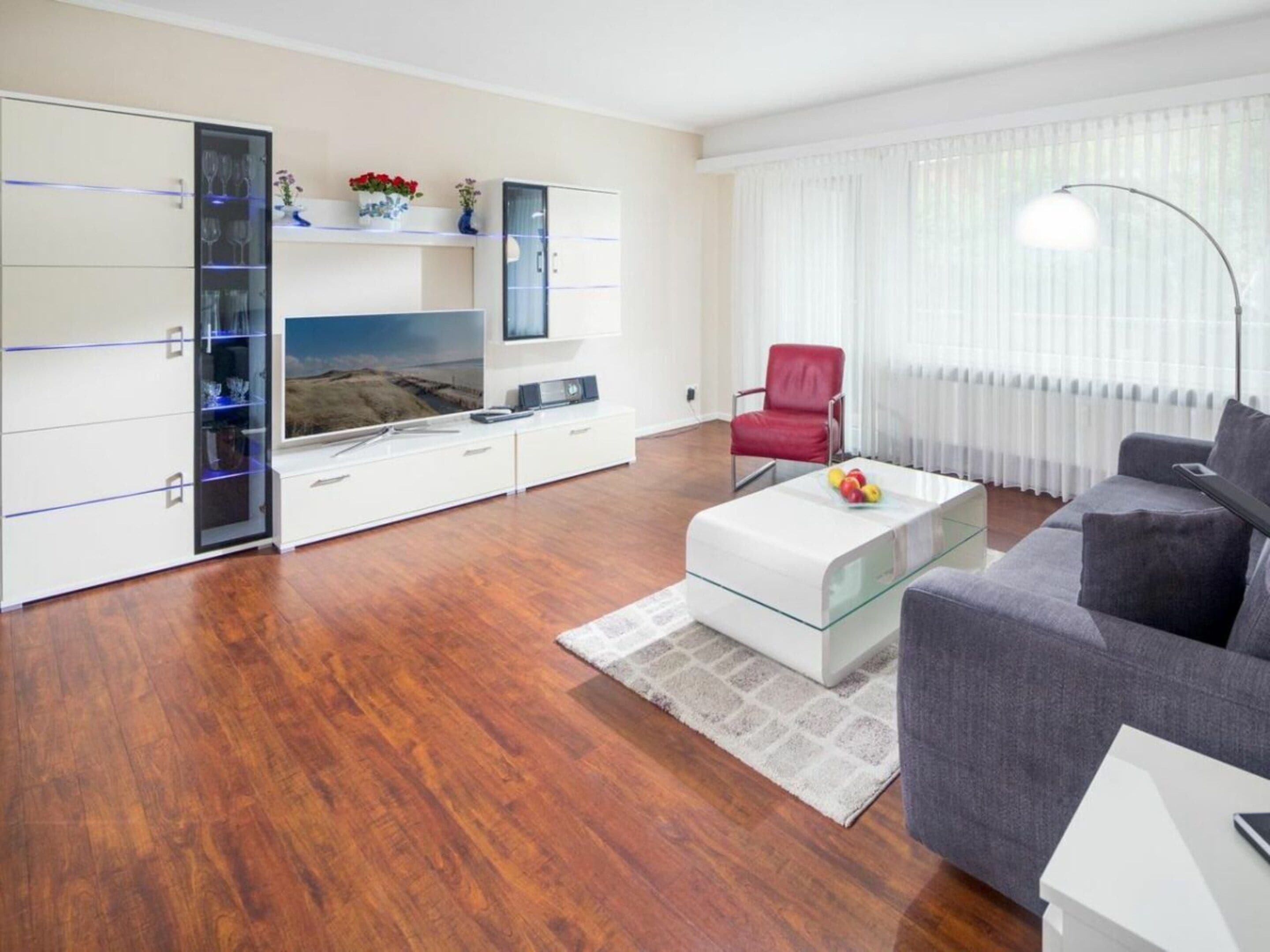 Ein modernes Wohnzimmer mit Fernseher, einem roten Sessel, einem grauen Sofa und einem weißen Couchtisch.