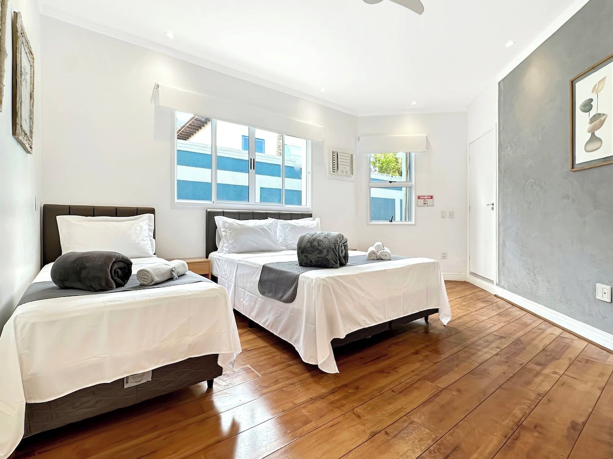 Quarto de hotel com duas camas de solteiro, piso de madeira, ventilador de teto e janelas amplas.