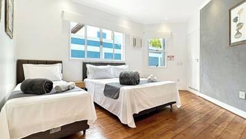 Quarto de hotel com duas camas de solteiro, piso de madeira, ventilador de teto e janelas amplas.