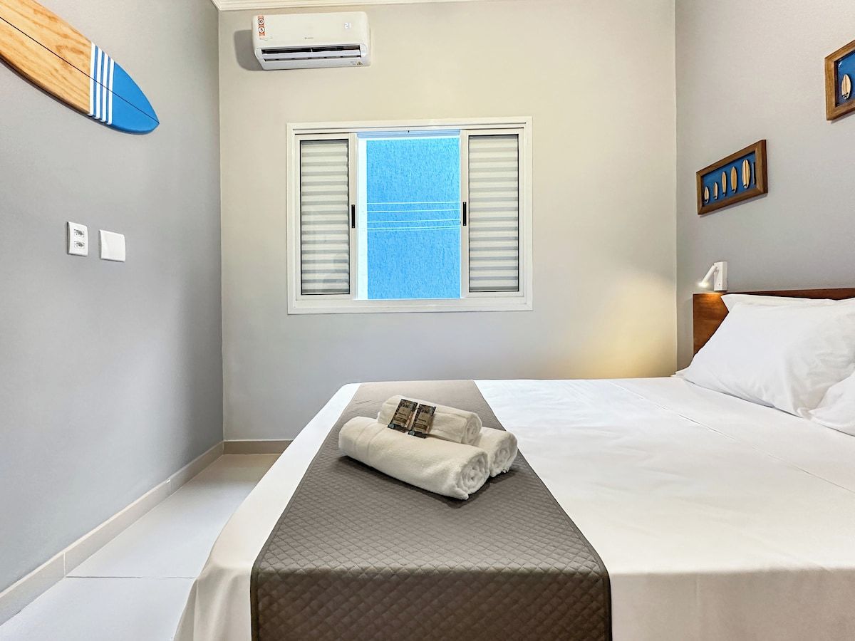 A hotel room with a bed, a window with shutters, an air conditioning unit, and a surfboard on the wall.