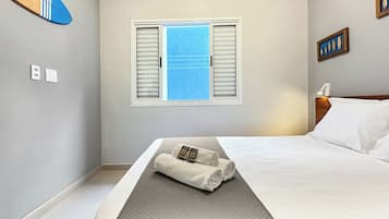 A hotel room with a bed, a window with shutters, an air conditioning unit, and a surfboard on the wall.