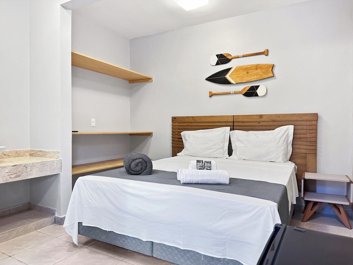 Quarto de hotel com cama, cabeceira de madeira, prateleira na parede e mesa de cabeceira pequena.