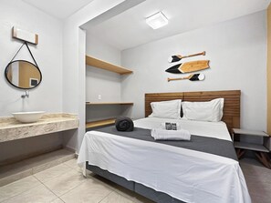 Habitación de hotel con cama, mesita de noche, espejo, estante y una tabla de surf fijada a la pared.