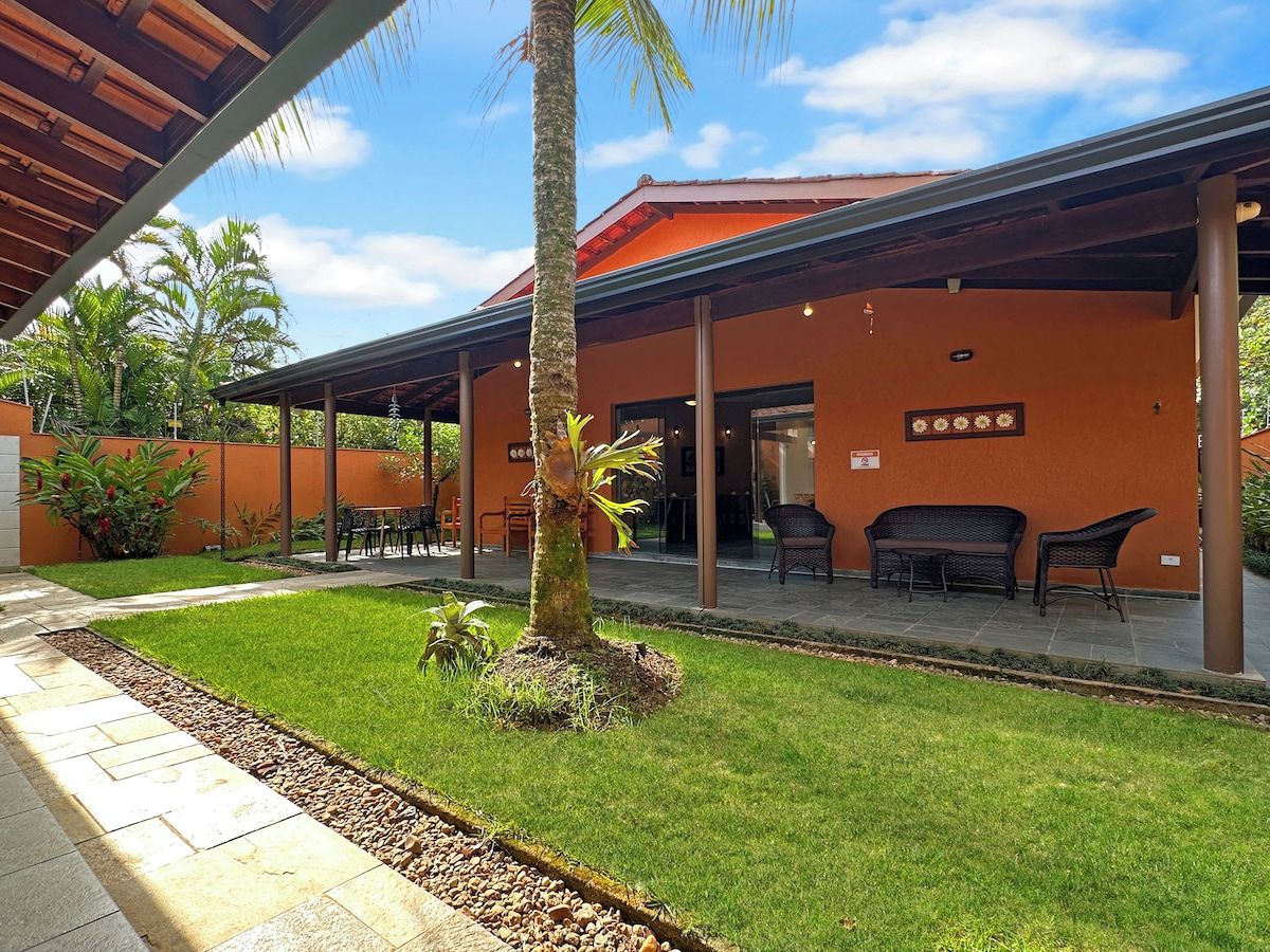 Uma área de estar coberta ao ar livre, com uma palmeira, um edifício de fachada laranja e céu claro.