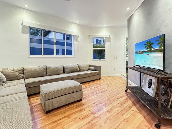 A living room with a sectional sofa, ottoman, wooden flooring, and a TV on a stand.