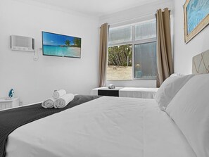 A hotel room with a bed, a TV displaying a beach scene, a window with curtains, and a wall-mounted air conditioner. - Maraka_12 · Pousada Maraka , Quarto 12 (Guarujá)