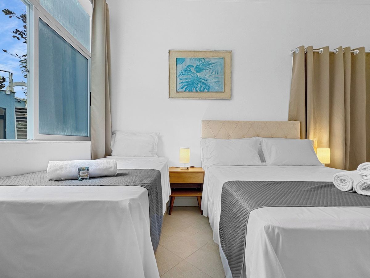 A hotel room with two beds, a nightstand with a lamp, a window with curtains, and a framed picture on the wall.