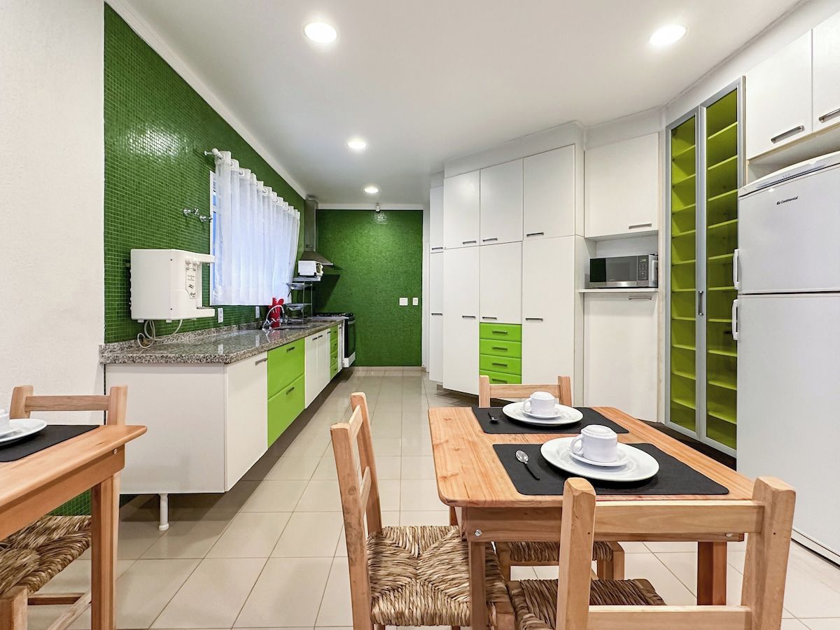 A modern kitchen with white cabinets, a green accent wall, a dining table with chairs, and a built-in microwave.