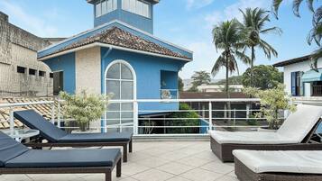 A rooftop terrace with lounge chairs, a small building with a blue facade, and palm trees in the background.