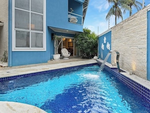 A swimming pool with a water feature, a chair, and a potted plant.