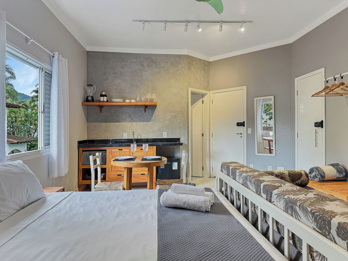A modern bedroom with a large bed, a sofa bed, a kitchenette, and a view of the outdoors.