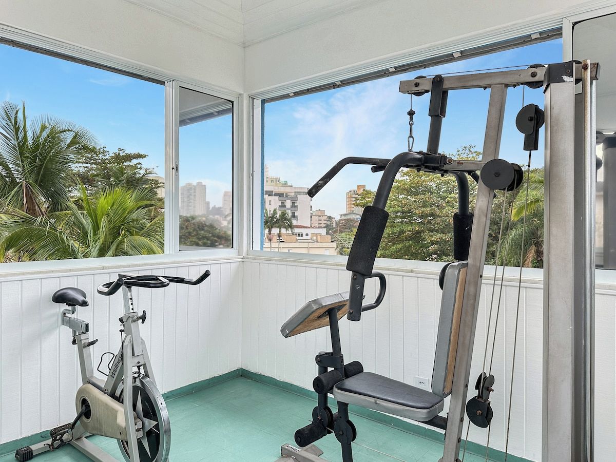 A gym with exercise equipment and a stationary bike, with a view of palm trees and buildings through large windows.