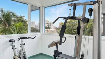 A gym with exercise equipment and a stationary bike, with a view of palm trees and buildings through large windows.