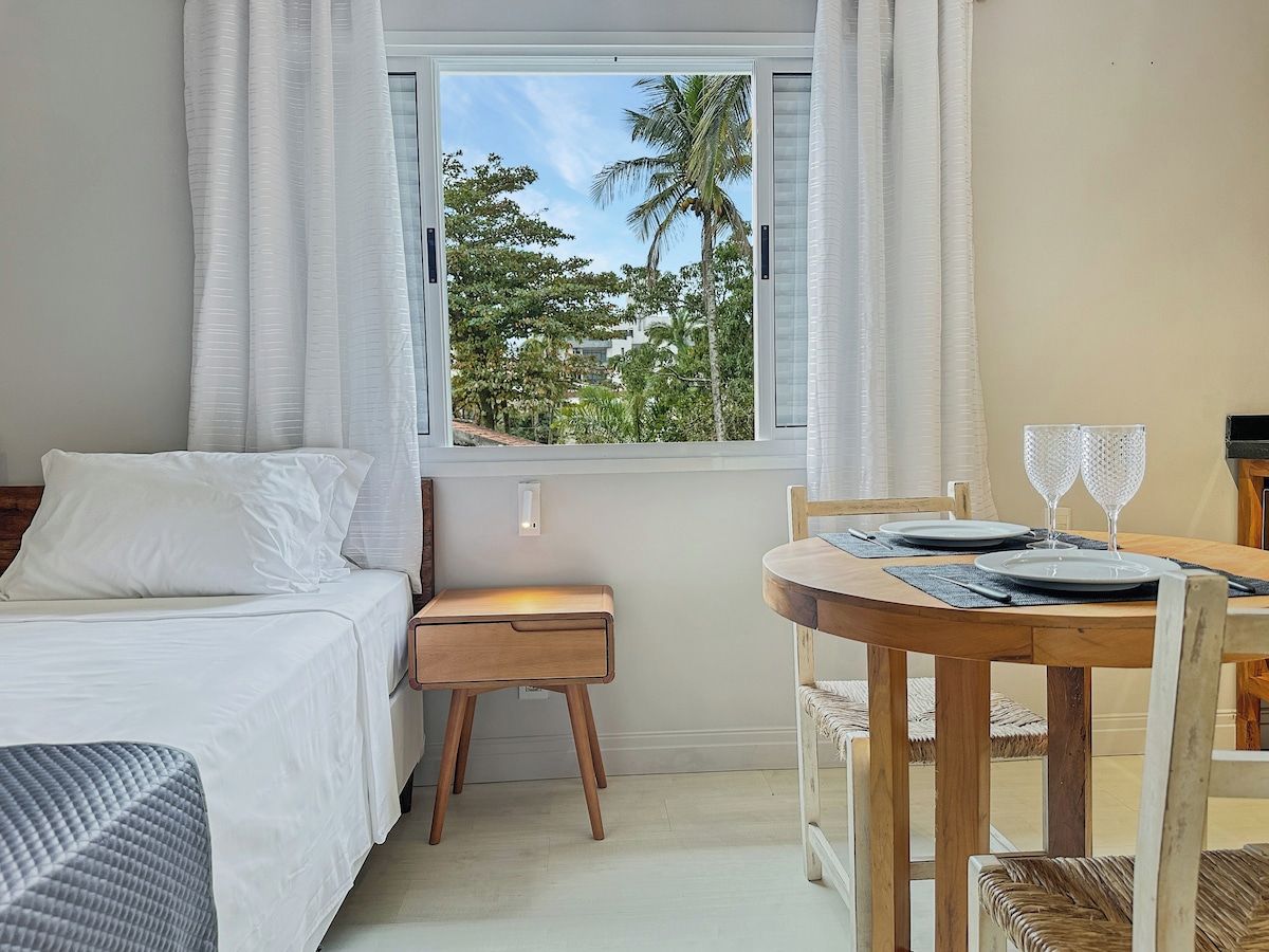 A hotel room with a bed, a small table with two glasses, and a window with a view of trees.