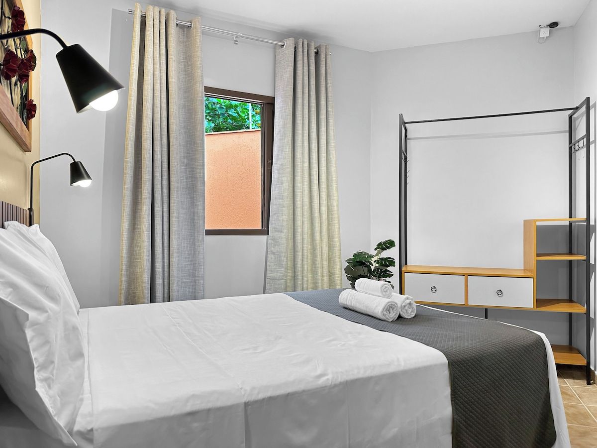 Habitación de hotel con cama, mesita de noche, espejo, estante y ventana con cortinas.